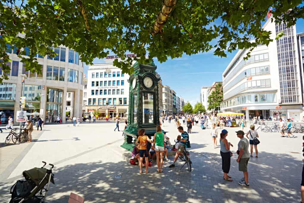Blick auf den Kröpcke in Hannover mit Menschen, Bäumen und modernen Gebäuden.