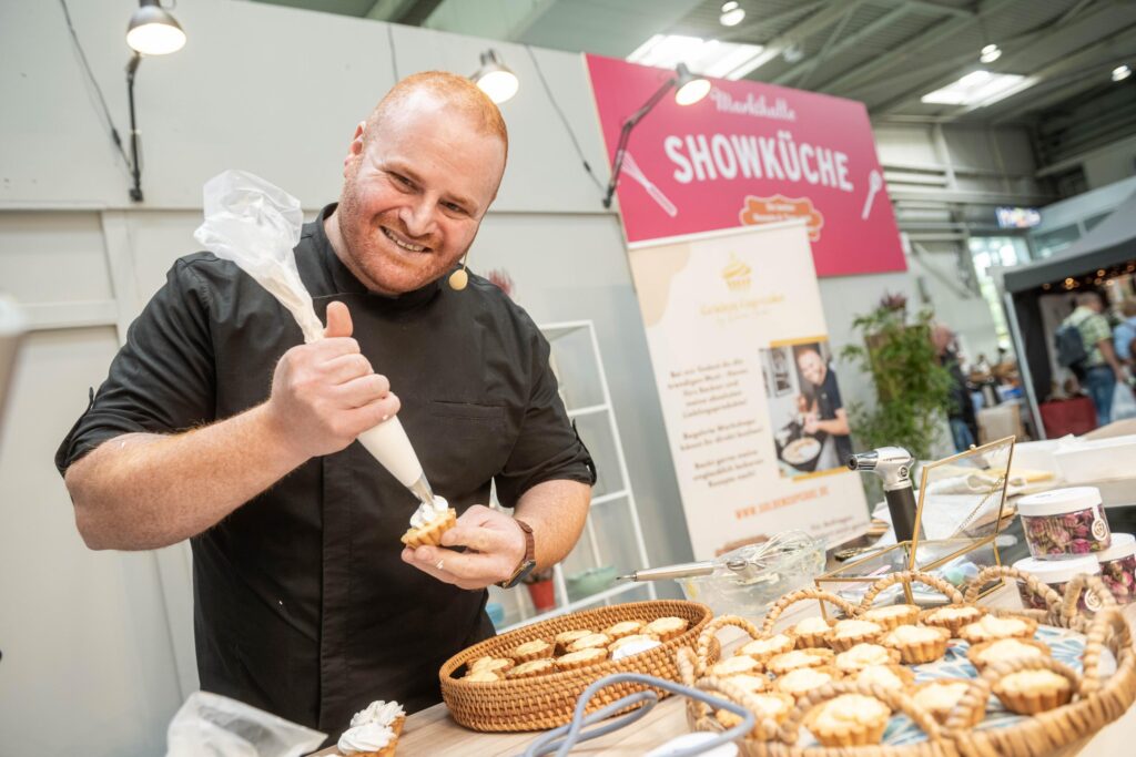 Ein Mann dekoriert Cupcakes. Er steht an einem Messestand der infa.