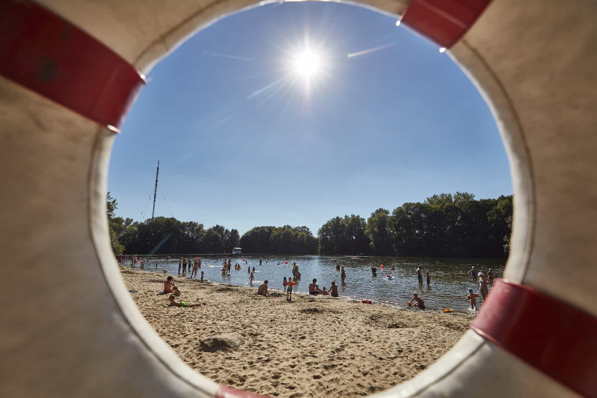 Blick durch eine Rettungsringöffnung auf das Strandbad Hemmingen mit Sonnenlicht, Sandstrand und Bad.