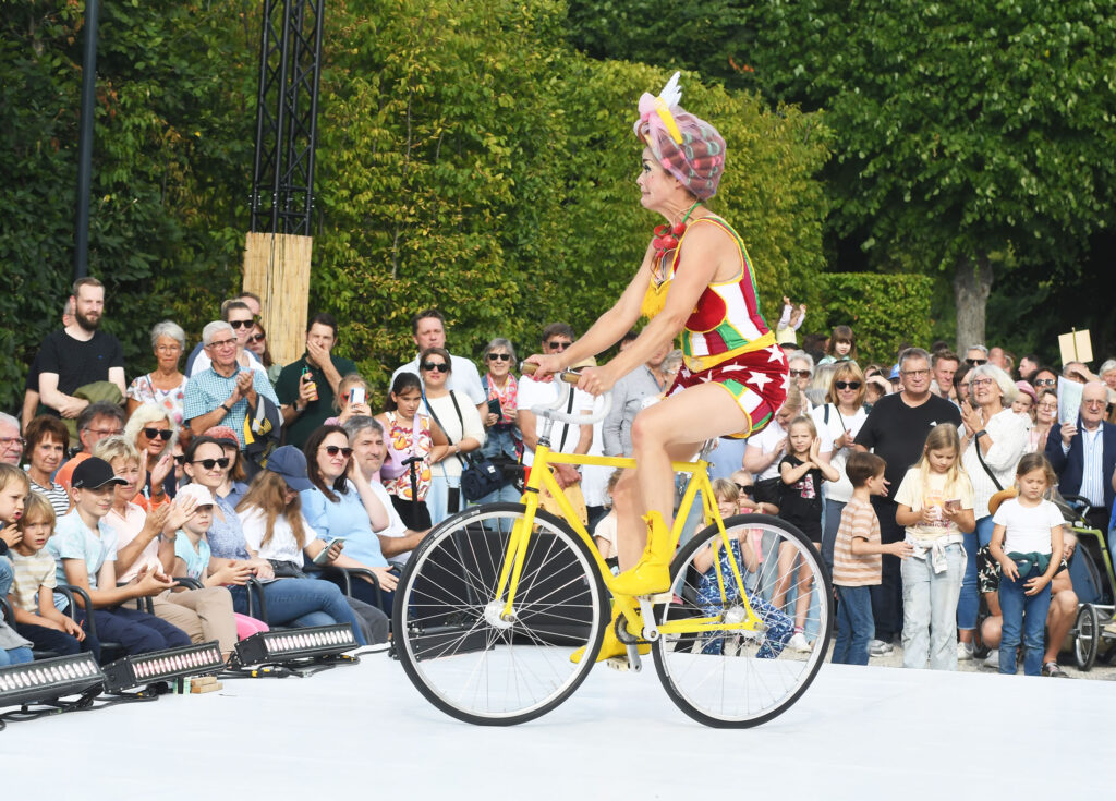 Künstlerin beim Kleinen Fest auf der Bühne auf einem gelben Fahrrad. Im Hintergrund sind Zuschauer zu sehen.