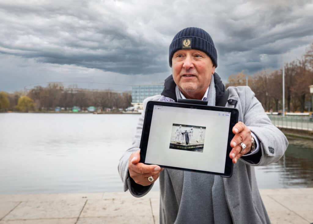 Mann zeigt Tablet mit Tatort-Foto am Wasser in Hannover bei bewölktem Himmel.