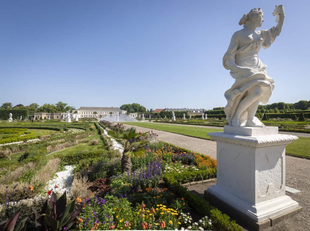 Blick auf den Herrenhausen Garten mit Skulptur und blühenden Blumen.