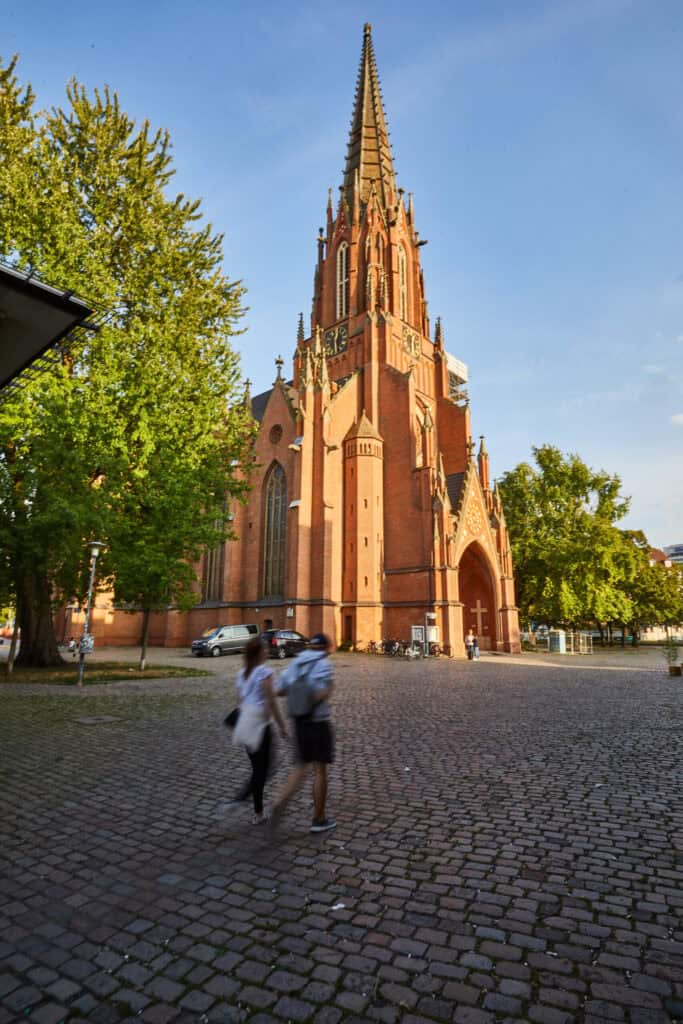 Historische Kirche in Hannover Nordstadt, umgeben von Bäumen, bei sonnigem Wetter, mit Spaziergänger.