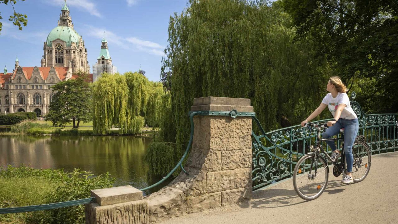 Mulher a andar de bicicleta junto à água com vista para a histórica Câmara Municipal de Hanôver.